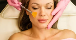 Woman receives a Chemical Peel in Kansas City, MO, on a spa chair, with pink-gloved hands applying product to her cheek using a small brush.